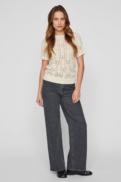 Pull écru à col rond de VILA®, avec un motif ouvert et associé à des jeans noirs et des chaussures.