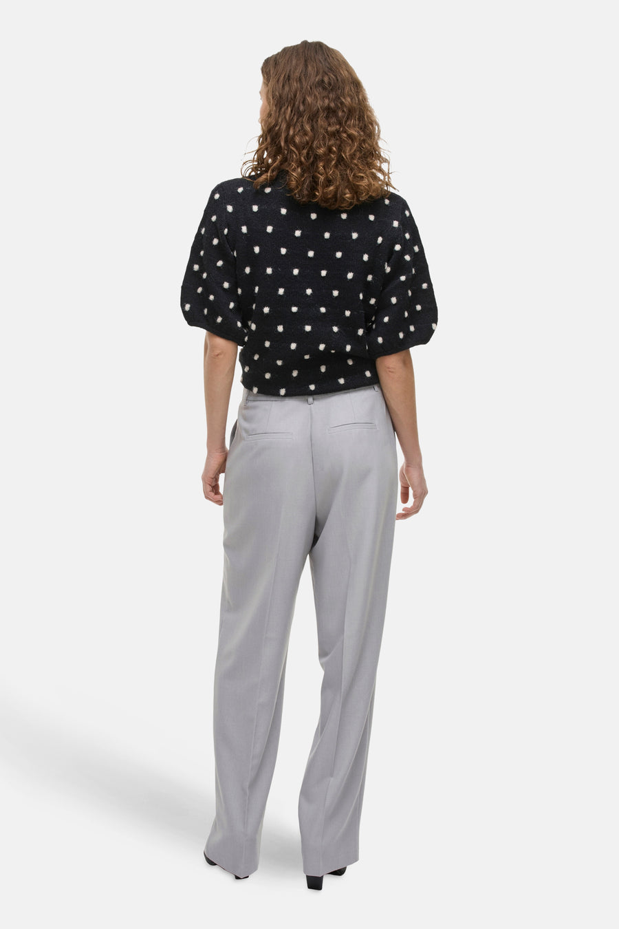 Achterkant van een vrouw met krullend haar in een zwarte trui met witte stippen en grijze pantalon.