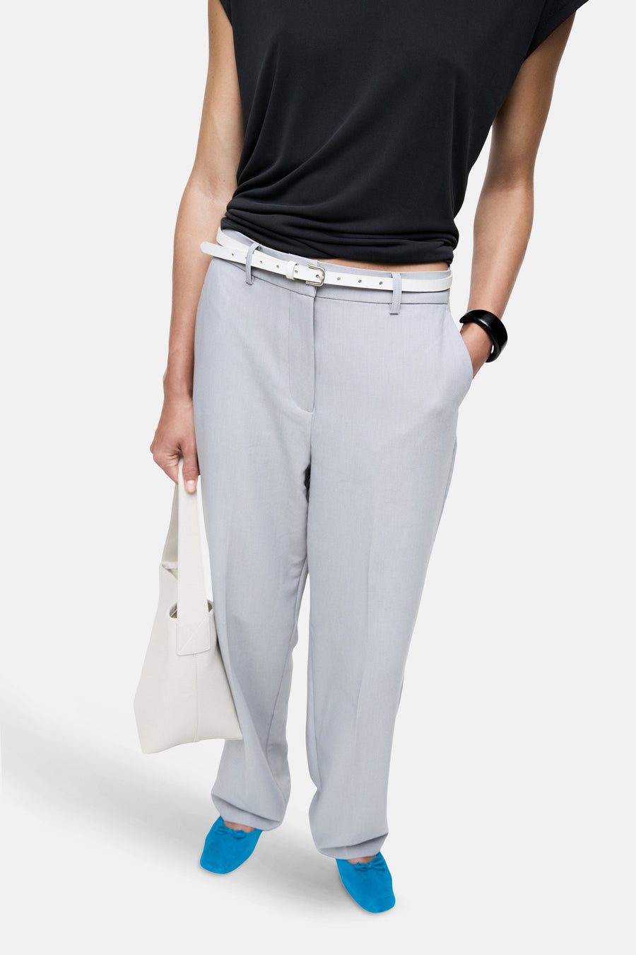 Een vrouwelijk model draagt een zwarte top, grijze pantalon met een witte riem, blauwe schoenen, een witte tas en een zwarte armband.