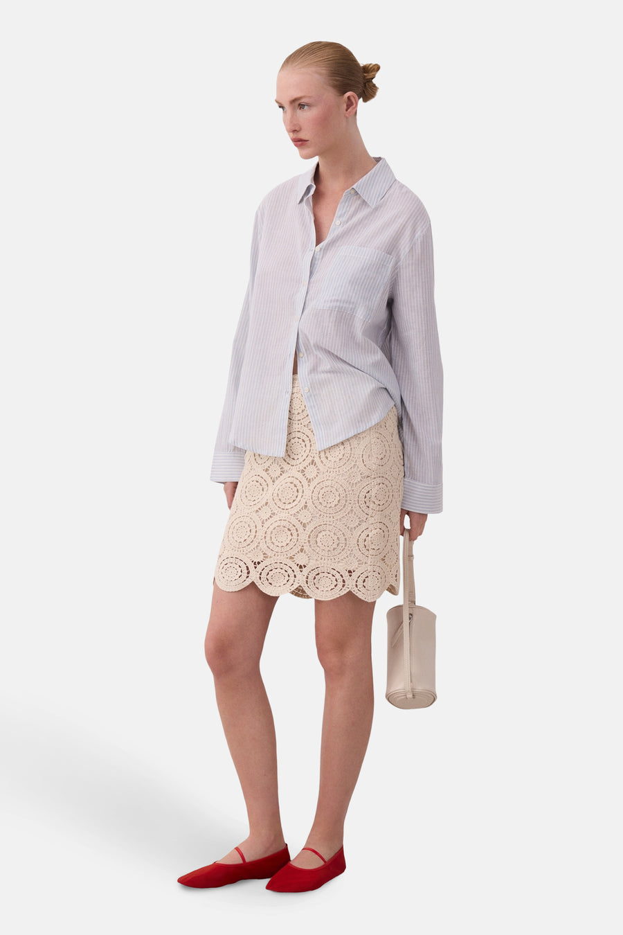 Vrouw in blauw gestreept overhemd, beige gehaakte rok, rode schoenen en beige handtas.