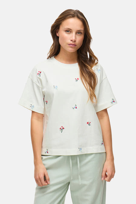 T-shirt blanc à manches courtes de VILA®, avec un motif brodé de fleurs, porté avec un short de jogging vert.
