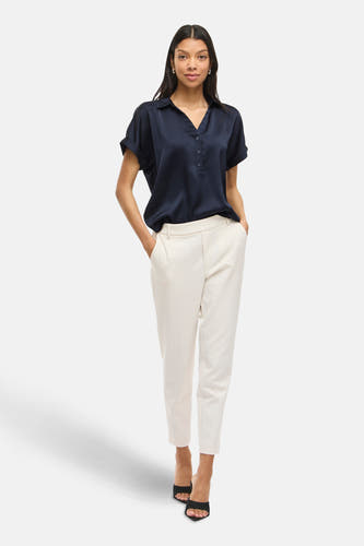 Blouse bleue à manches courtes de VILA, avec une fermeture à boutons et portée avec un pantalon blanc.
