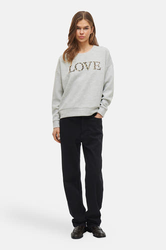 Grijze sweater van VILA, met "LOVE" print in luipaardmotief, gecombineerd met donkere broek en leren schoenen.
