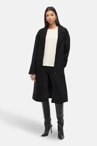 Zwarte lange jas van VILA, met knoopsluiting, gedragen met een witte top, zwarte jeans en hoge laarzen.
