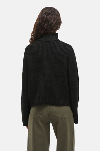 Achterkant van een vrouw met een zwarte coltrui en groene broek.