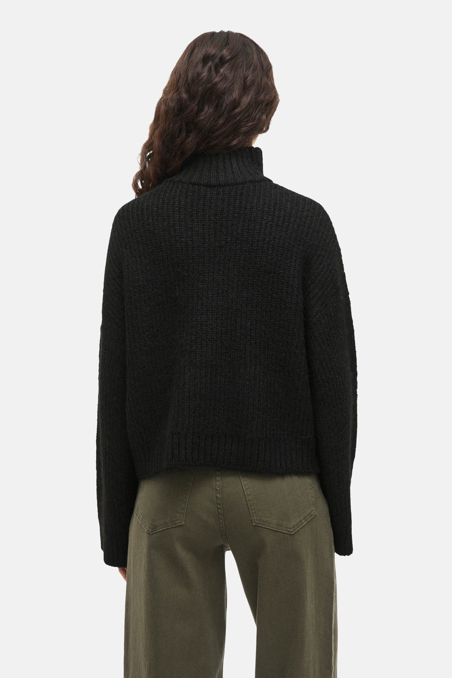 Achterkant van een vrouw met een zwarte coltrui en groene broek.