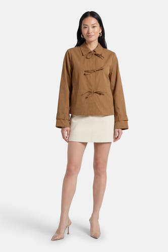 Beige korte jas van VILA® met knoopdetail en lange mouwen, gecombineerd met een witte rok en nude pumps.
