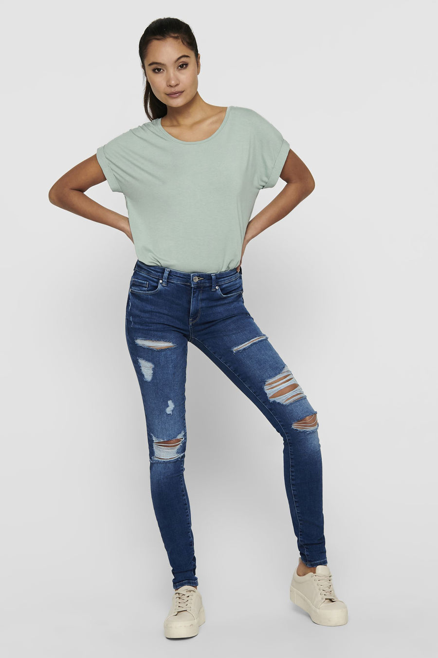 Vrouw met groene top en gescheurde jeans, staand.

