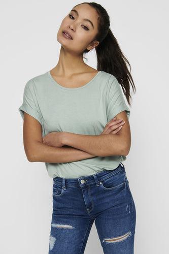 Femme avec un t-shirt vert et des jeans bleus, les bras croisés.