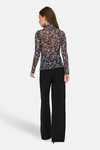 Vrouw in camouflage shirt met lange mouwen en zwarte, wijde broek, staand van achteren.