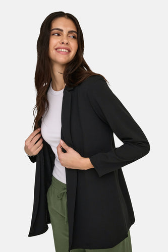Femme avec un blazer noir, une chemise blanche et un pantalon vert, souriant.