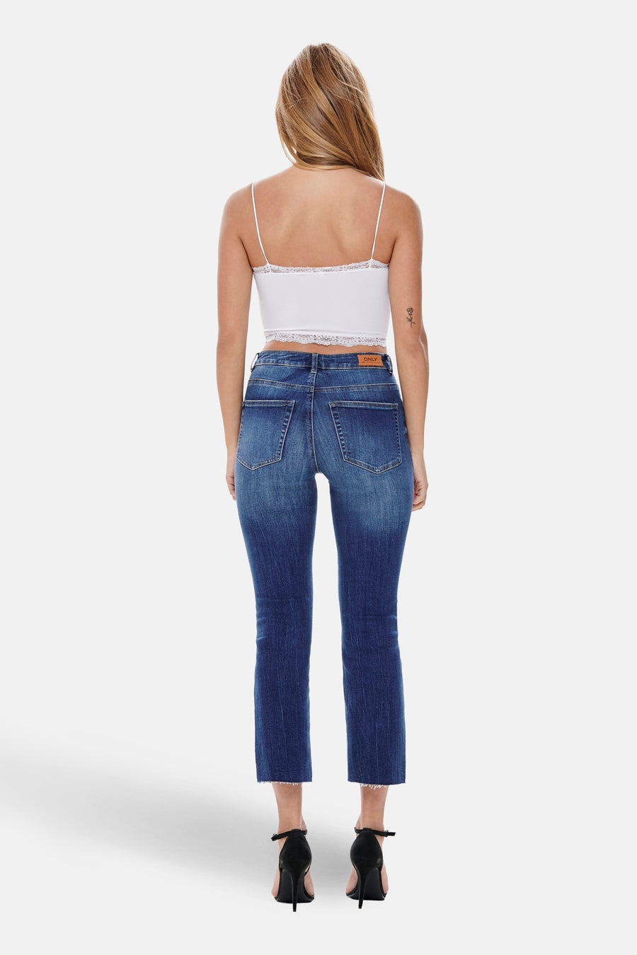 Achterkant van een vrouw met een witte top en blauwe jeans, staand op zwarte hakken tegen een witte achtergrond.