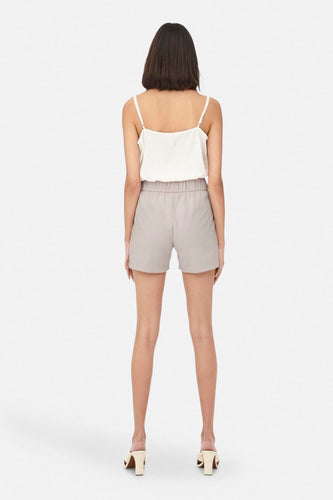 Dos d'une femme en une chemise blanche avec des bandes de spaghetti et des shorts gris avec une ceinture élastique, debout sur des talons.