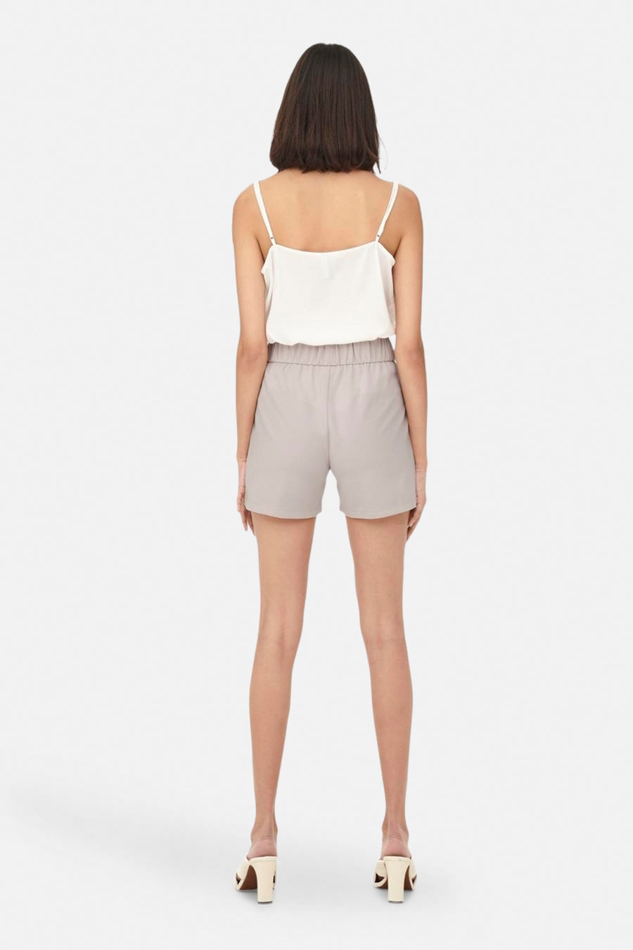 Dos d'une femme en une chemise blanche avec des bandes de spaghetti et des shorts gris avec une ceinture élastique, debout sur des talons.