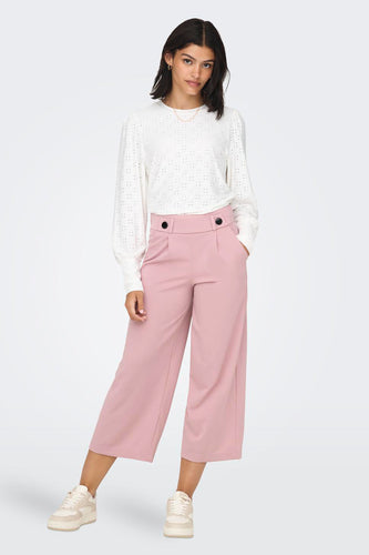 Vrouw in witte blouse en roze culottes, staand met handen in de zakken tegen een witte achtergrond.