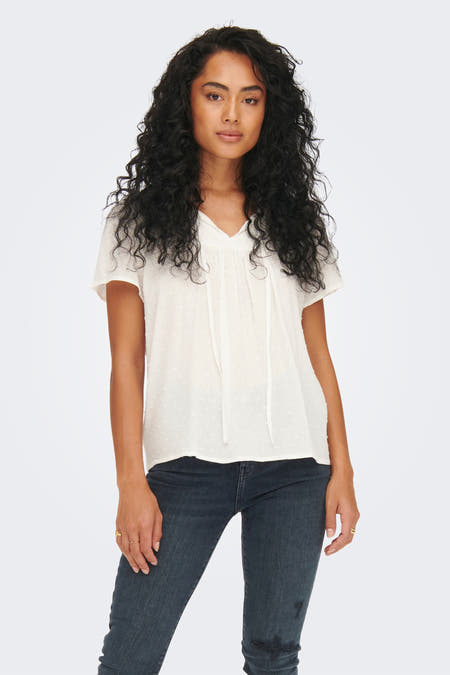 Blouse blanche à manches courtes de JDY, avec un col en V et des cordons, portée avec un jean foncé.