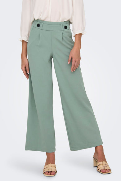 Pantalon de costume vert deY JDY, deses larges et une taille haute avec des boutons décoratifs, associé à une blouse blanche et des sandales à talon.