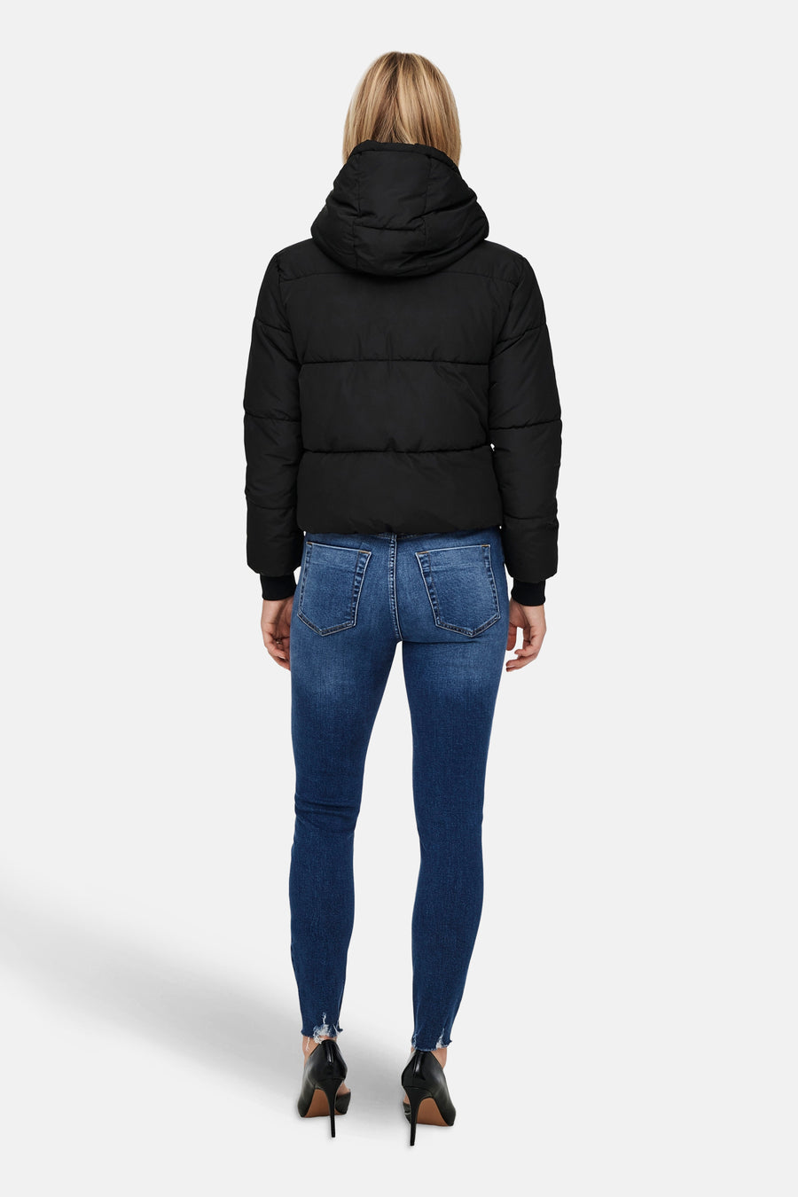 Dos d'une femme en un manteau de fourrure noir, des jeans bleus et des talons noirs sur un fond blanc.