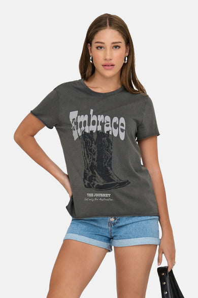 T-shirt gris à manches courtes de ONLY, avec un imprimé de bottes de cowboy et le texte "Embrace", porté avec des shorts en denim.