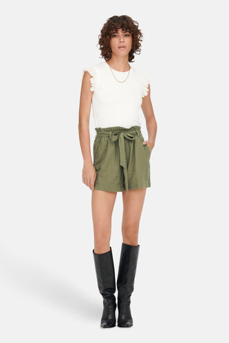 Vrouw in witte top, groene shorts en zwarte laarzen staand.