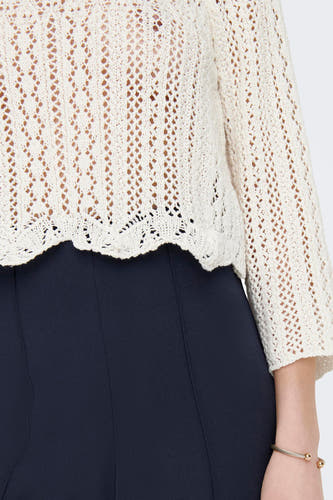 Pull écru d'ONLY, avec motif ajouré, combiné avec un pantalon bleu foncé et un bracelet en or.