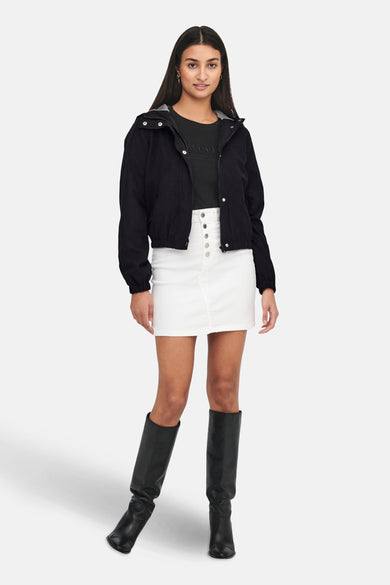 Manteau court noir de JDY, avec capuche, associé à une jupe blanche et des bottes noires.