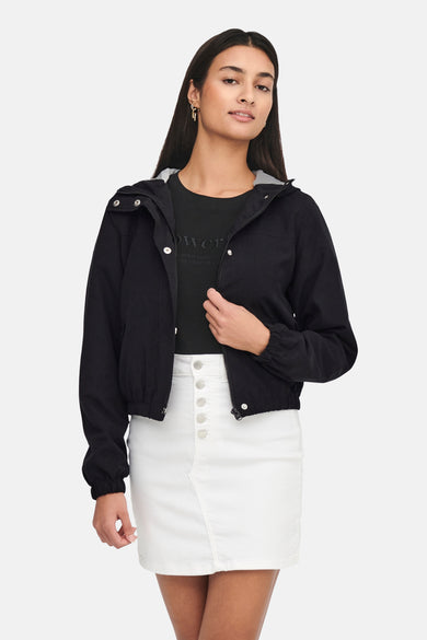 Manteau court noir de JDY, avec capuche et porté avec une jupe blanche et un T-shirt noir.