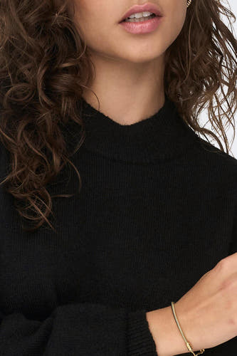 Robe noire courte de JDY, avec manches longues, un col haut et un bracelet en or subtil.
