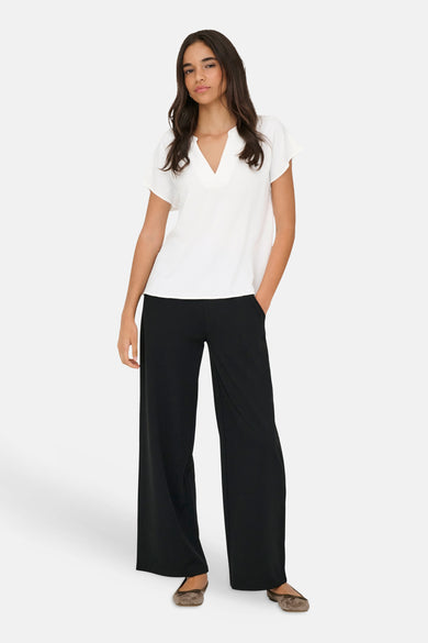 Chemisier blanc à manches courtes de JDY, avec col fendu et texture subtile. Porté avec un pantalon noir ample et des ballerines taupe.