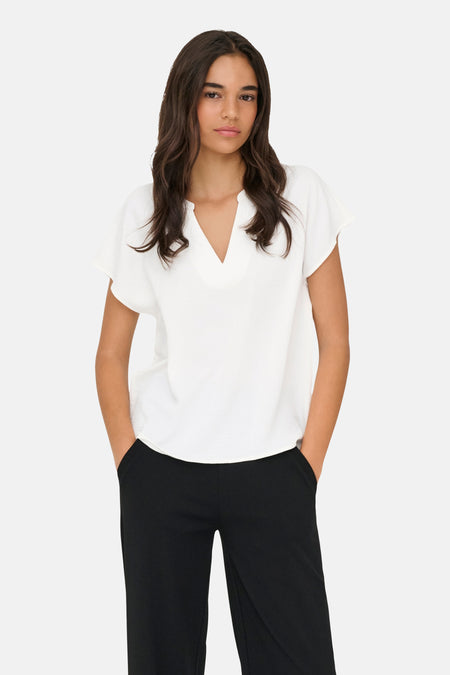 Chemisier blanc de JDY avec col en V, manches courtes amples et tissu texturé, porté avec un pantalon noir.