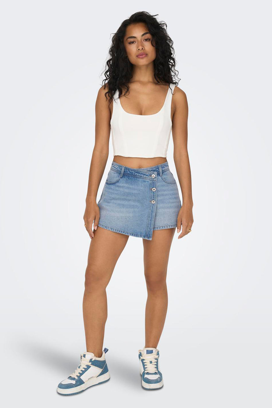 Short de denim bleu clair ONLY, avec une fermeture à bouton asymétrique et porté avec un haut blanc et des baskets hautes.