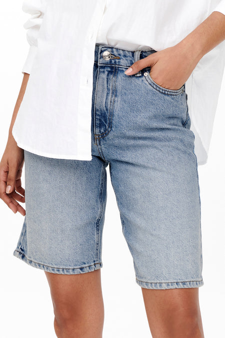 Shorts en denim bleu clair de ONLY, avec une main dans la poche et un chemisier blanc.