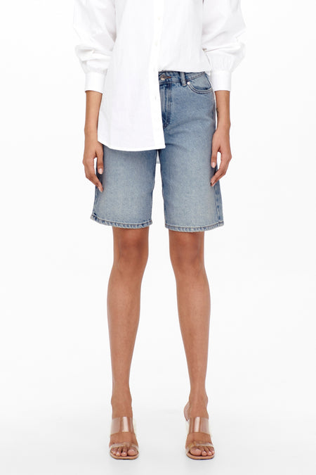 Courtes Bermudas denim bleu clair d'ONLY, portées avec un chemisier blanc et des sandales transparentes.