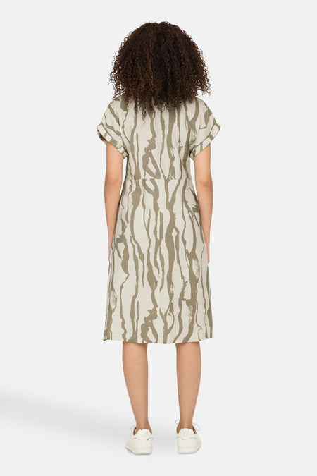 Femme avec des cheveux bouclés arrièrerière, robe robe une beige et un vert olive motif, baskets blanches, fond blanc.