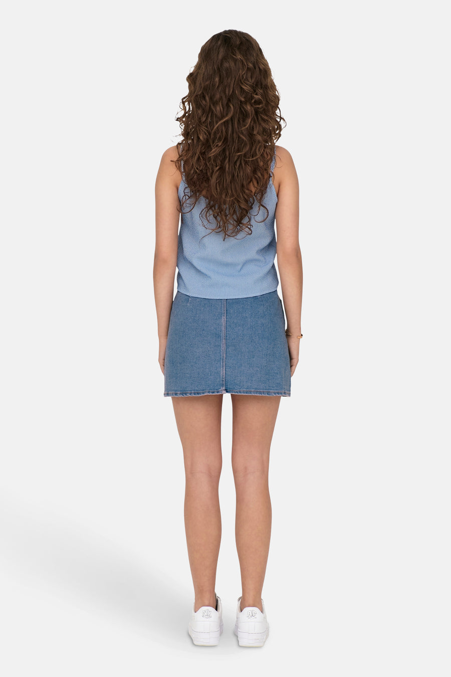 Vrouw, achterkant, krullend haar, blauw-wit gestreepte top, denim rok, witte sneakers.