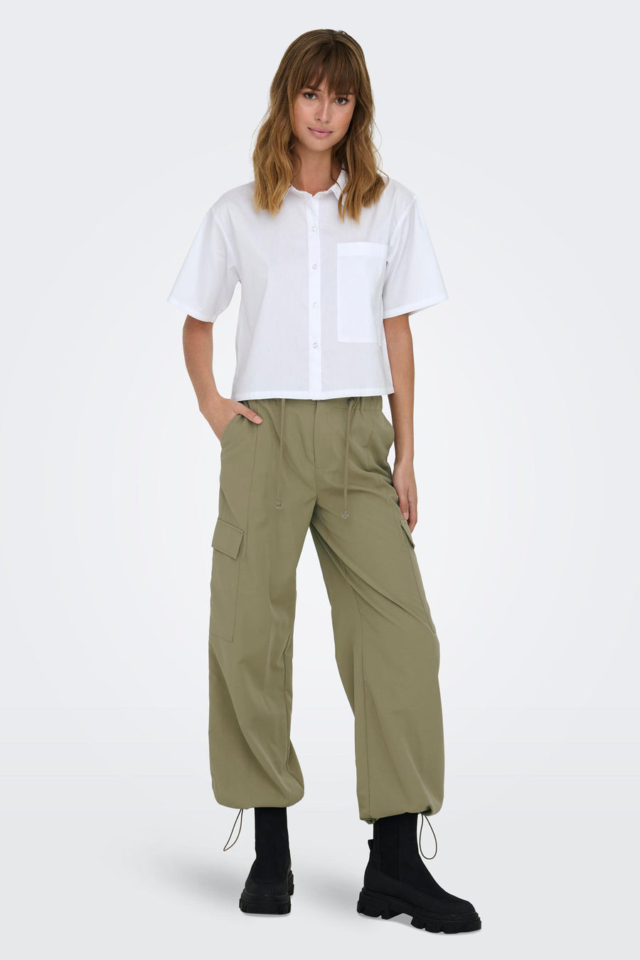 Pantalon de travail vert ONLY, avec une ceinture à la taille et porté avec un chemisier blanc et des bottes noires.