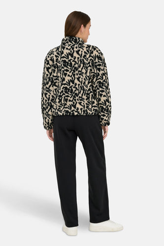 Achterkant van een vrouw in een beige en zwarte fleece jas met zwarte broek en witte schoenen op een witte achtergrond.