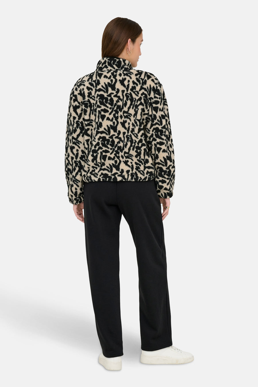 Achterkant van een vrouw in een beige en zwarte fleece jas met zwarte broek en witte schoenen op een witte achtergrond.