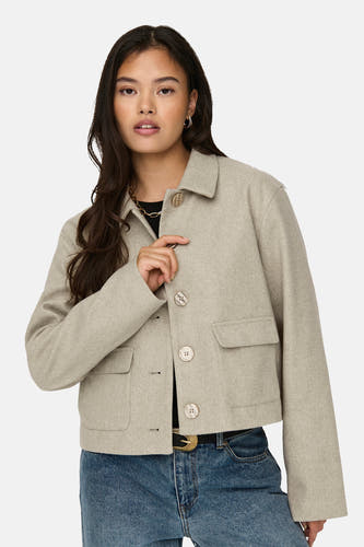 Manteau court beige de JDY, avec fermeture à boutons, poches et porté avec une ceinture noire et des jeans bleus.