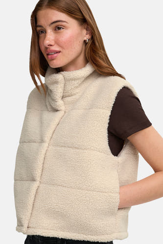 Doudoune sans manches couleur crème en tissu de peau de mouton avec col montant, de la marque ONLY®, portée sur un pull côtelé marron foncé, vue de profil.