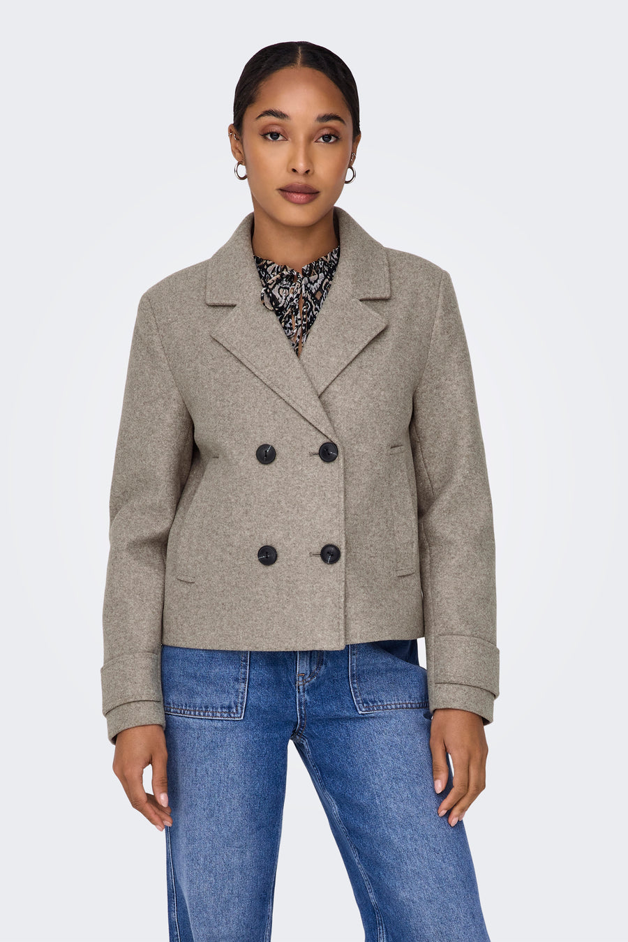 Manteau court beige d'ONLY, avec des boutons doubles et porté avec une blouse imprimée et un jeans bleu.