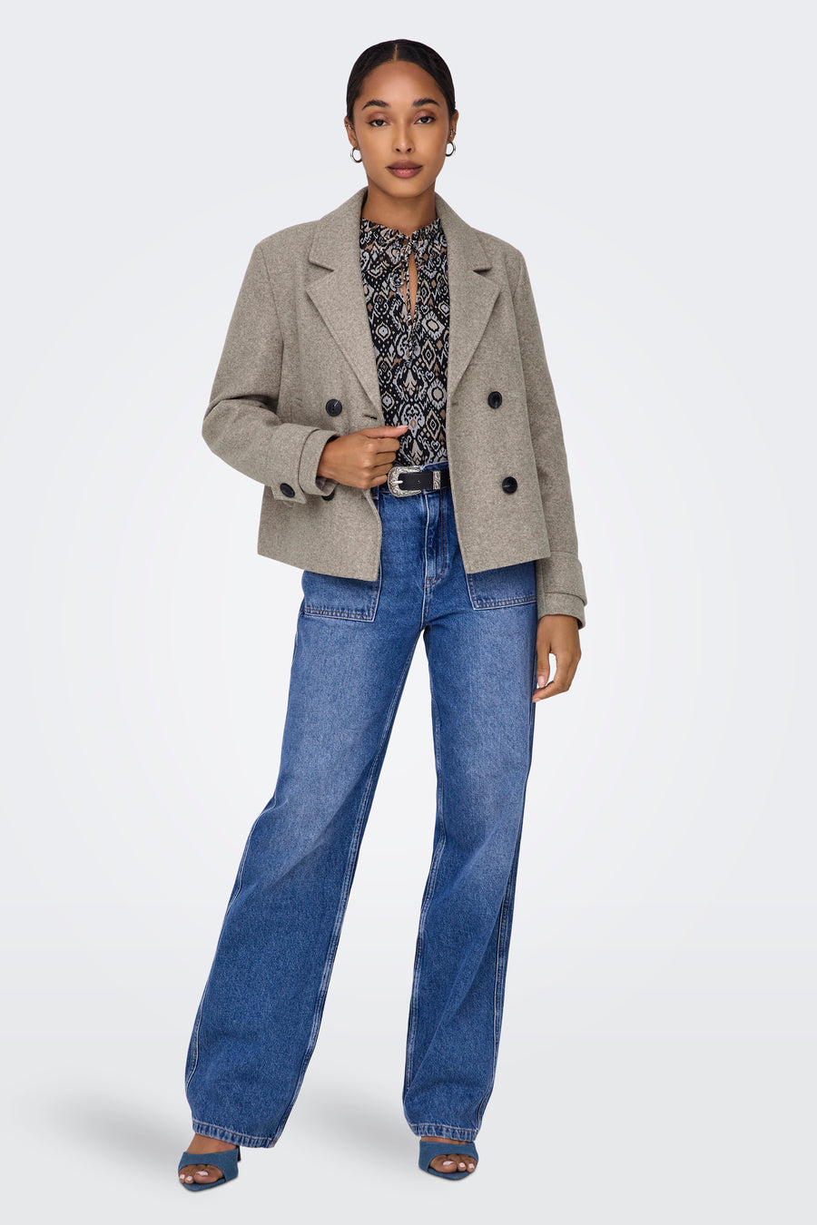 Manteau court beige d'ONLY, avec une fermeture à double bouton, porté sur une blouse imprimée et des jeans larges.