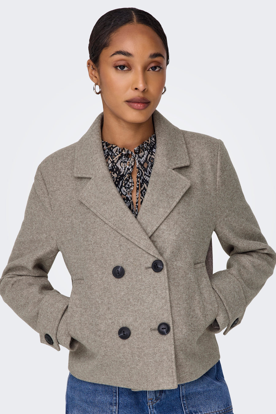 Manteau court beige d'ONLY, avec des boutons noirs et une blouse à fleurs en dessous, porté avec un jeans bleu.