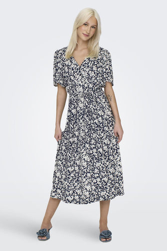 Vrouw in bloemige midi-jurk met blauwe schoenen op witte achtergrond.