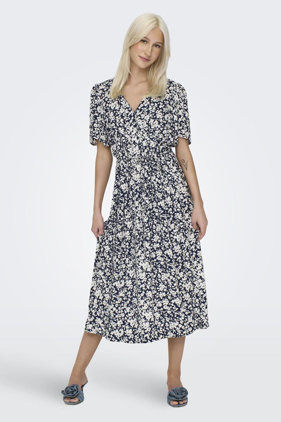 Vrouw in bloemige midi-jurk met blauwe schoenen op witte achtergrond.