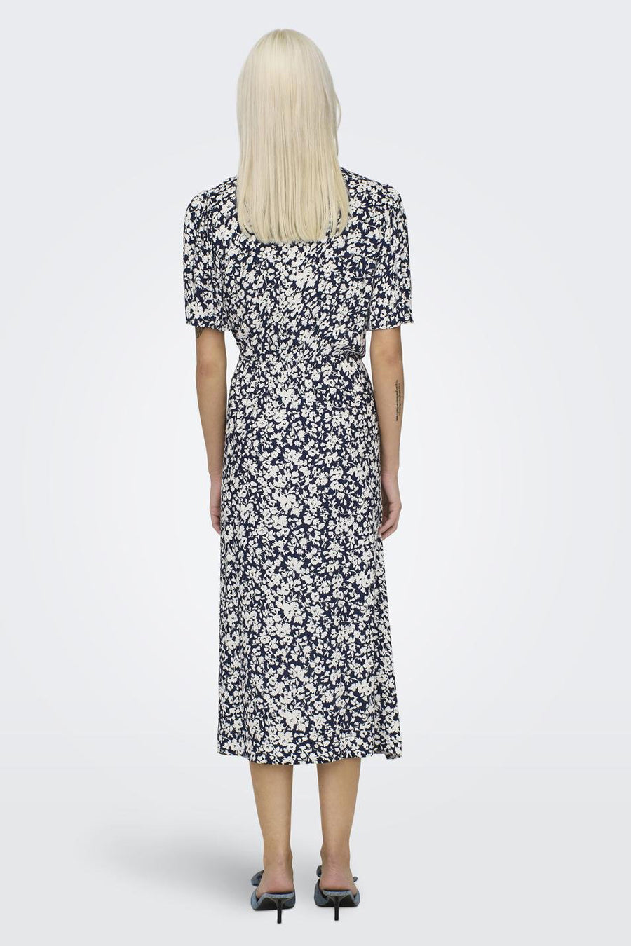 Achterkant van een vrouw in een marineblauwe bloemenjurk met korte mouwen en zwarte hakken op een witte achtergrond.