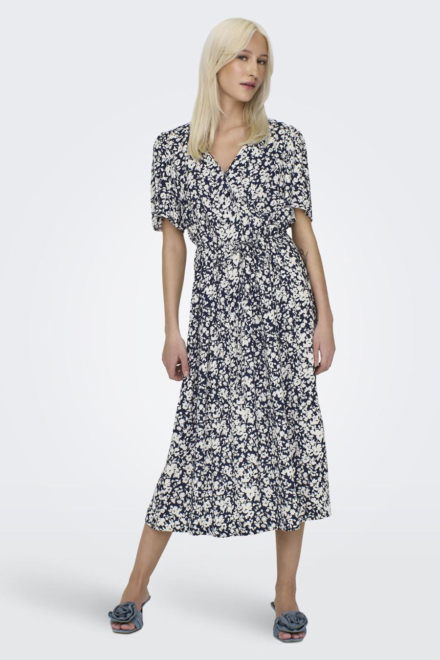 Een vrouw draagt een blauw-witte bloemenjurk en blauwe muiltjes.