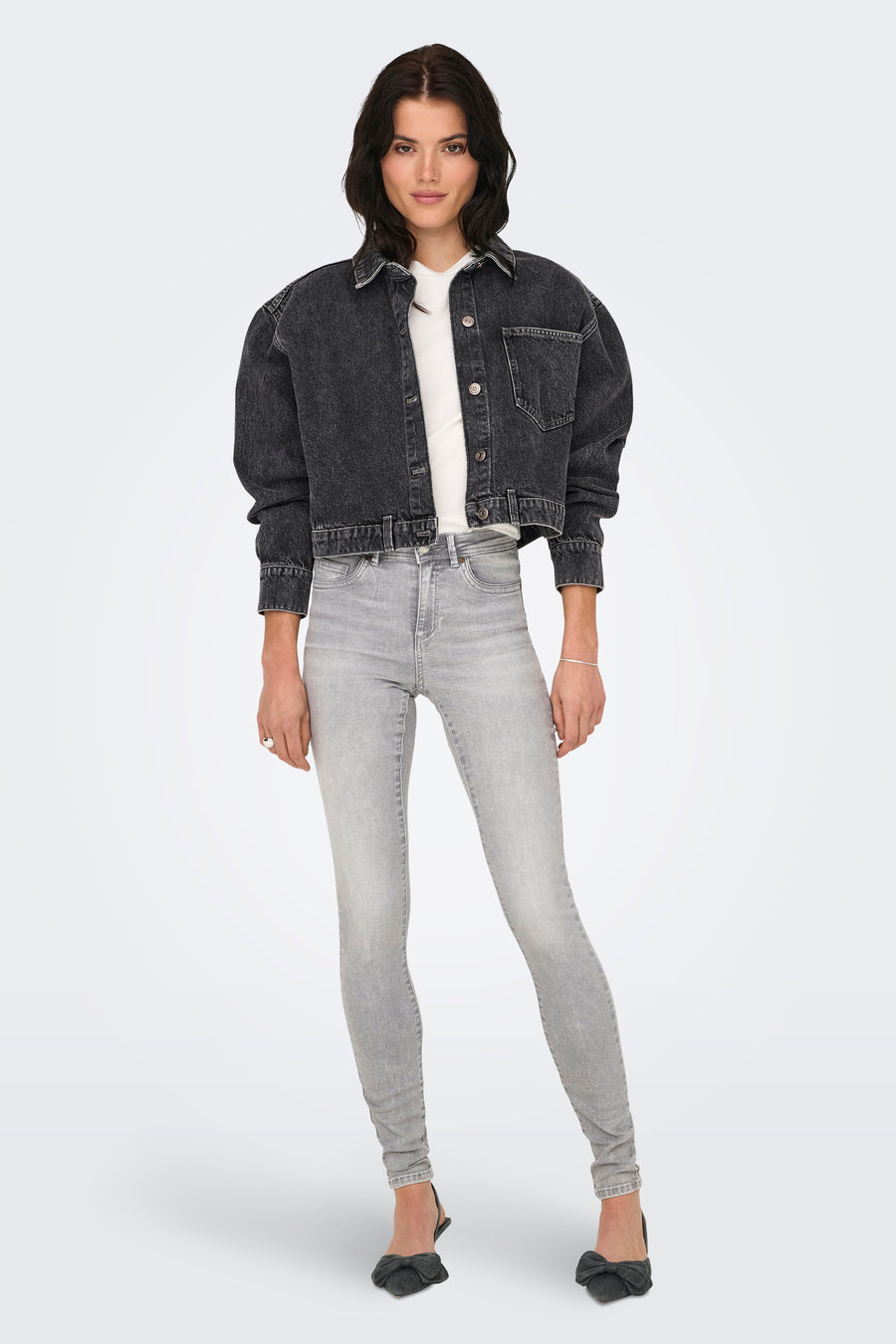 Donkergrijze denim jeansjas van ONLY, gedragen over een wit t-shirt met lichtgrijze skinny jeans en zwarte hakken met strik.

