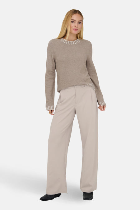 Vrouw met blonde haren in een bruine trui en beige pantalon, poserend voor een witte achtergrond.