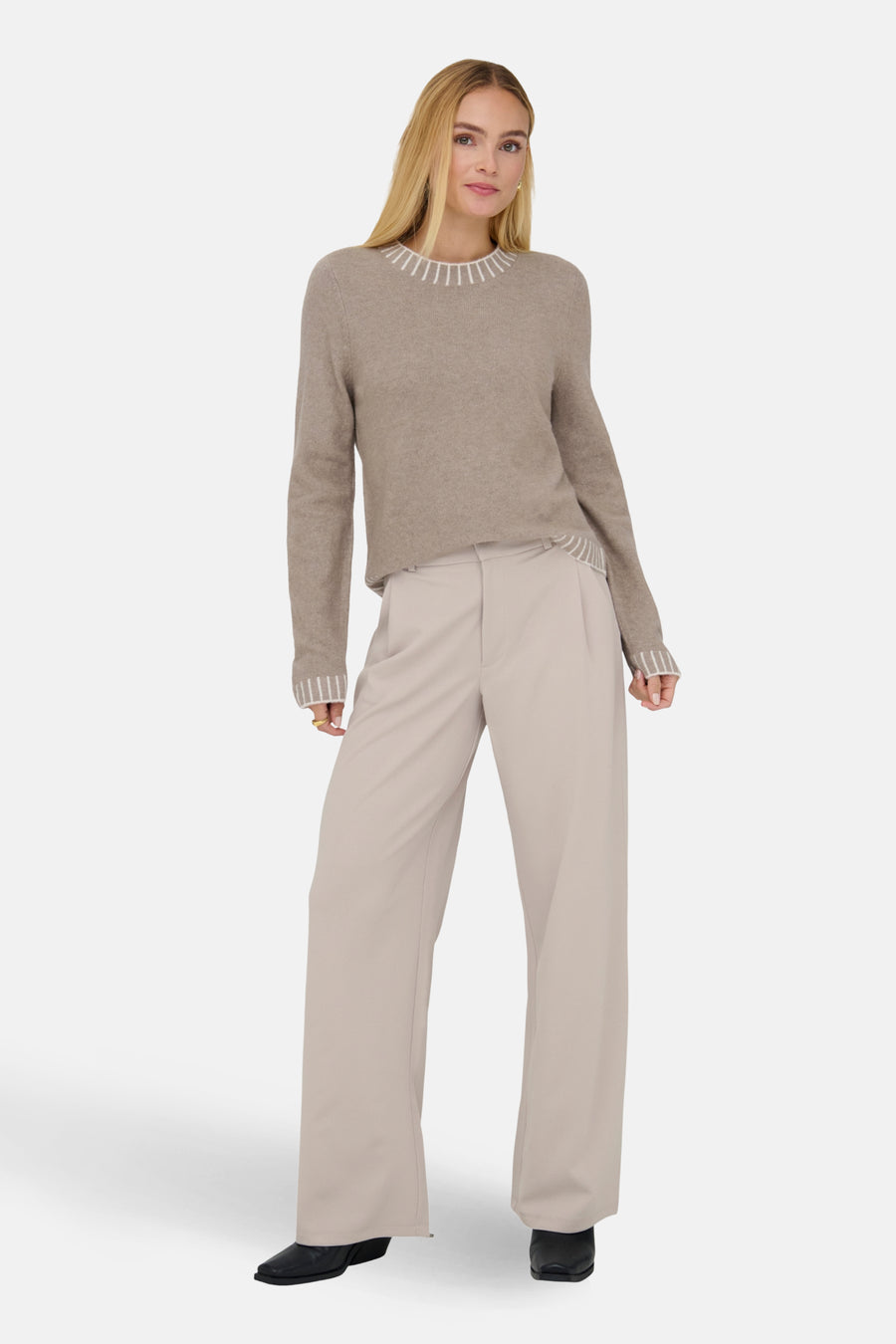 Vrouw met blonde haren in een bruine trui en beige pantalon, poserend voor een witte achtergrond.